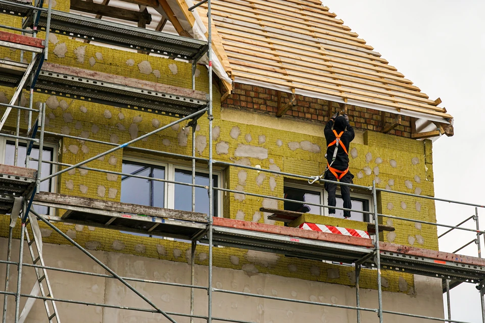 Photographie d'un artisan appliquant l'isolation thermique par l'extérieur (ITE) : l'ouvrier en équipement de sécurité (gilet, gants) appose une plaque isolante en polystyrène ou laine minérale sur la façade d'une maison avec un mortier-colle, montrant clairement le système d'isolation en couches superposées (isolant, enduit de base, armature, finition). La scène se déroule en extérieur sur un chantier de rénovation résidentielle, avec échafaudage visible, illustrant professionnellement un travaux d'isolation thermique RGE.