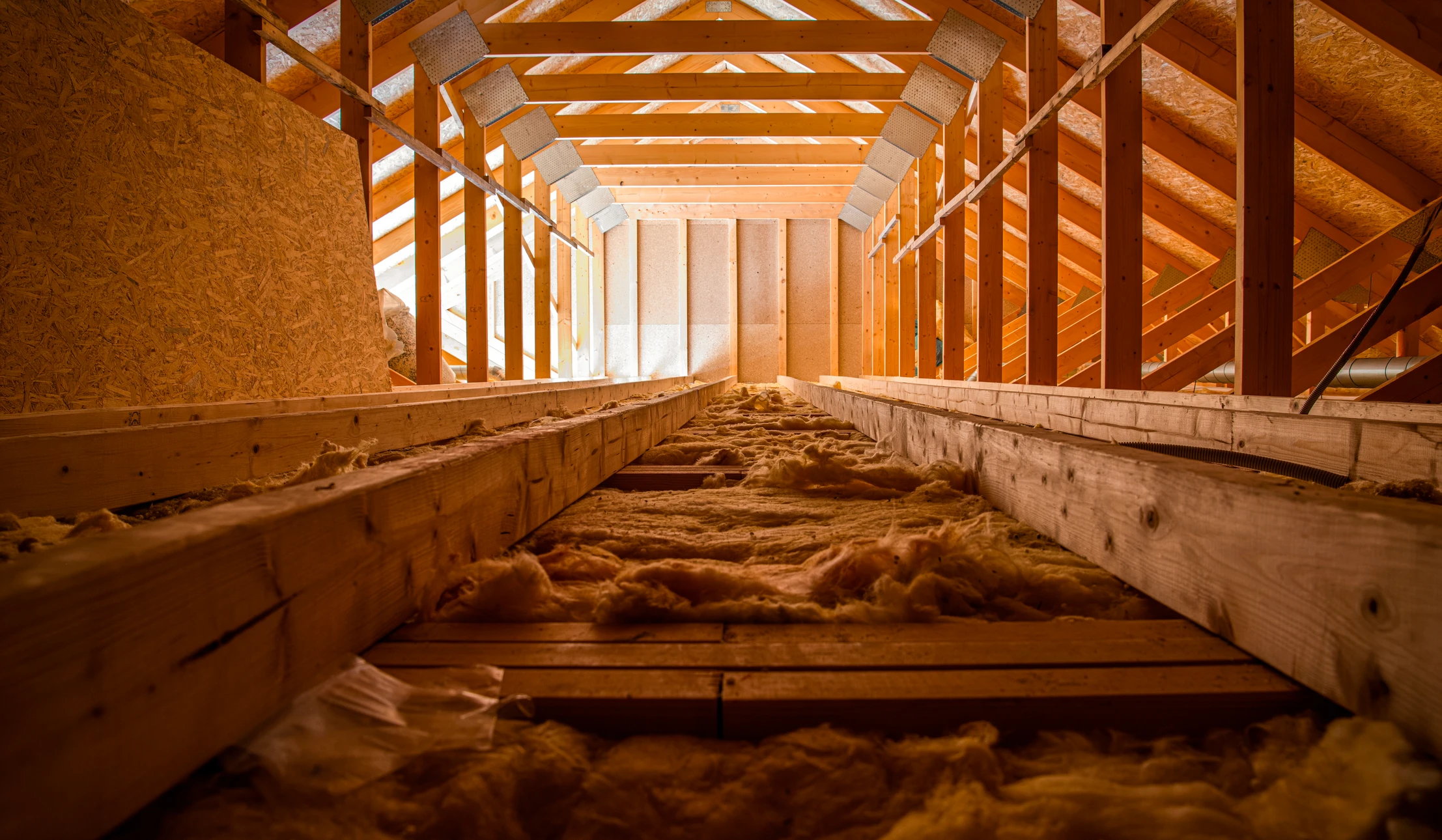 Photographie professionnelle en couleur d'une personne ou d'une équipe en tenue de chantier (combinaison, casque de sécurité, gants) effectuant les travaux d'isolation dans des combles : déroulement de matériau isolant (laine minérale, ouate de cellulose) entre les chevrons d'une charpente, sous l'éclairage naturel d'une lucarne. L'accent doit être mis sur la technique d'installation, la qualité du travail et l'environnement de chantier authentique.