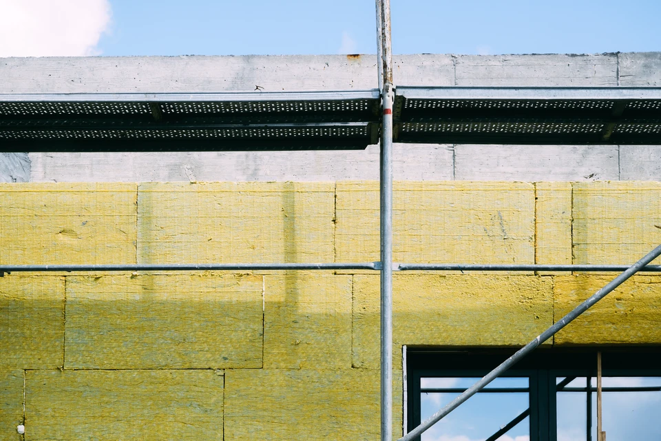 Photographie réaliste d'une maison individuelle en cours de rénovation avec isolation thermique par l'extérieur. La façade montre des panneaux isolants (polystyrène expansé ou laine minérale) de couleur blanche ou grise partiellement installés sur les murs, avec des ouvriers équipés (baudrier, casque) en train de poser les panneaux isolants ou l'armature de finition. La maison doit être un bâtiment résidentiel classique, et l'arrière-plan doit montrer clairement les travaux d'isolation en cours, illustrant comment la protection thermique se fait par l'extérieur sans modification de l'espace intérieur.